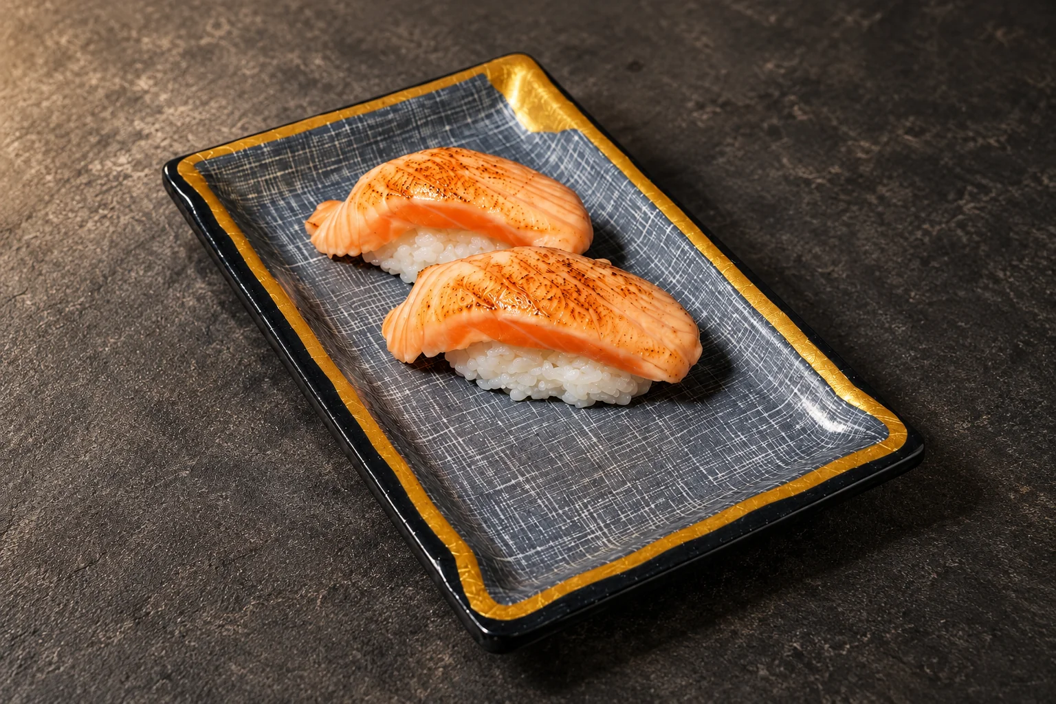 Two flamed salmon nigiri on a rectangular blue-grey plate with a gold rim.