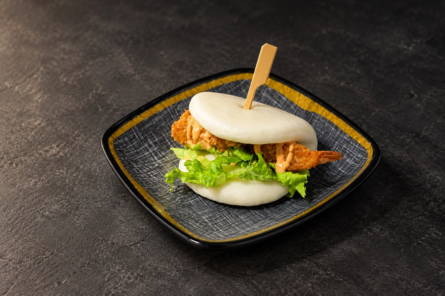 Bao bun filled with tempura shrimp and salad, on a square plate with a gold rim.